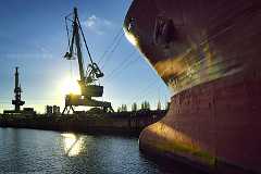 lange_leine Ein Frachtschiff liegt am Kai. Von seinem riesigen Bug führen mehrere Taue optisch zu einem Kran der sich als Shioluette vor der tief stehen Sonne abhebt.