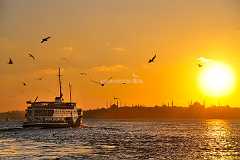 oriental-sundown Eine Fähre fährt kurz vor Sonnenuntergang von Kadiköy nach Eminönü auf dem Marmara Meer und wird von umkreisenden Möven begleitet. Im Hintergrund die...